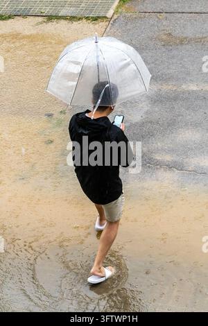 High-angle view of a person walking through a street covered in muddy floodwater during torrential rain, protected by a clear dome umbrella dotted wit Stock Photo