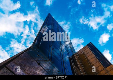 An upward view emphasizes the sharp edges of a modern office building clad in reflective glass and stone. White clouds appear both in the vivid blue s Stock Photo