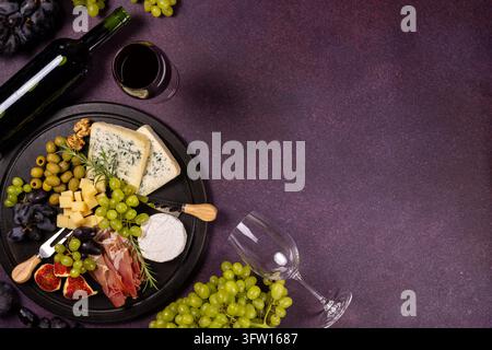 Charcuterie board presenting various cheeses, grapes, figs, olives, walnuts, prosciutto, and rosemary, served with a bottle of red wine and a full gla Stock Photo