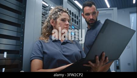 IT Specialists Review Server Performance on Laptop While Discussing Metrics and Adjusting LED Indicators, Demonstrating Collaboration, System Monitoring, Configuration, and Infrastructure Control. Stock Photo
