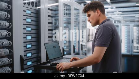 Male System Administrator Analyzes Server Telemetry on Laptop While Female Engineer Works on Nearby Racks, Combining Software Diagnostics with Physical Infrastructure Inspection inside Modern Data Center. Stock Photo