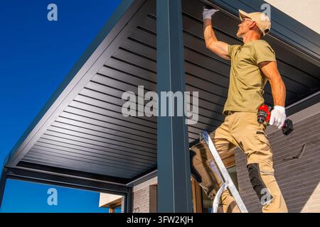 Low angle view of a worker on a stepladder using a cordless drill on the outer beam of a louvered pergola beside a house. Stock Photo