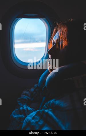 Woman with neck pillow resting beside airplane window at dusk, soft blue light on her face, Vietnam Stock Photo