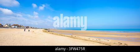Small figures walk along the wide sandy beach at low tide on Sword Beach, with coastal houses in the distance under a bright blue sky. Stock Photo