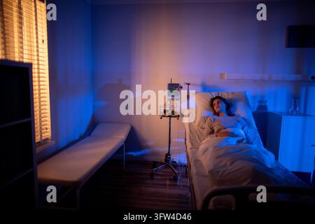 Hospital room wide view empty patient distant in frame, evening light through blinds casts shadow across bed and guest chair, mood of isolation and Stock Photo