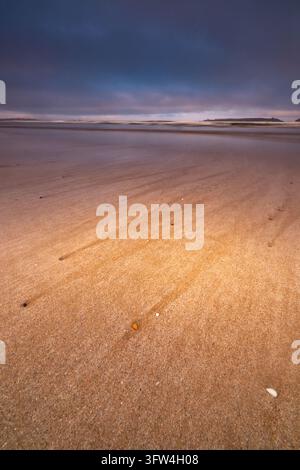 A low-angle vertical shot showing tiny stones on a vast flat sand shore as gentle tide water creates reflections under a cloudy purple evening dusk sk Stock Photo