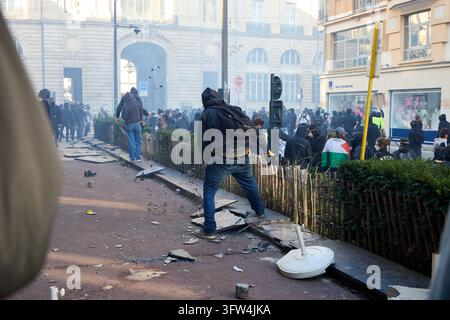 RENNES, FRANCE — OCTOBER 17, 2026: A hooded person with a backpack stands on a planter edge among scattered slate as a crowd gathers in smoke. Stock Photo