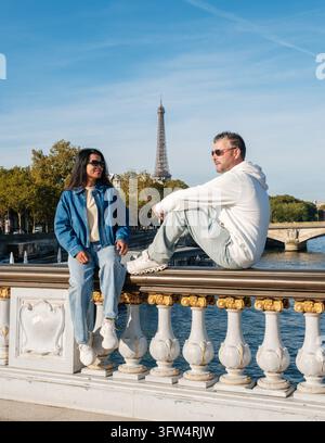A relaxed couple sits on the ornate Pont Alexandre III bridge above the Seine, with the Eiffel Tower in the background on a bright day in Paris, Franc Stock Photo
