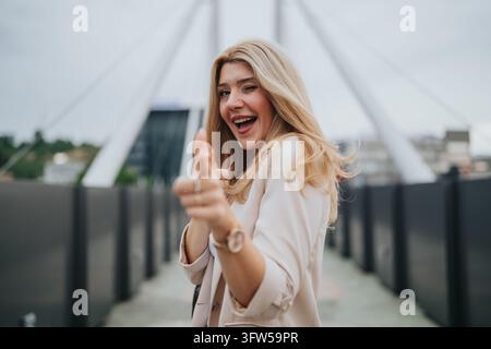 Smiling blonde woman in a light blazer points at the camera while winking on a pedestrian bridge. Stock Photo