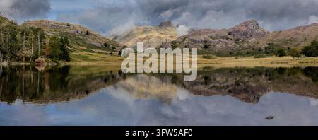 England, Cumbria, Autumn colors sweep across rugged mountains above a calm lake, where rippling water reflects the hills beneath a cloudy blue sky and Stock Photo