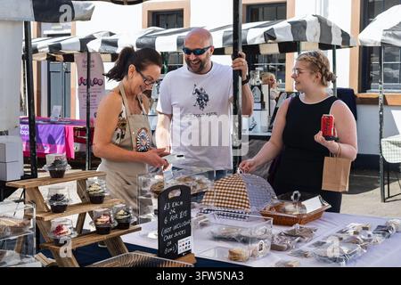 Ayr, UK - June 21, 2026: Woman wearing apron processes payment while bald man in U.S. Polo Assn. t-shirt and young woman browse baked goods under blac Stock Photo