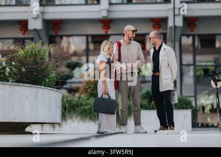 Three colleagues chat and smile while standing outside an office building. Casual business conversation among friends in an urban outdoor setting. Stock Photo