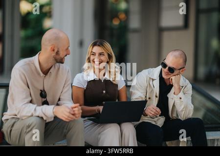 Three colleagues sit together outdoors, smiling and working on a laptop. The casual group chat captures friends collaborating in an informal outdoor m Stock Photo