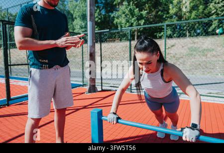 A woman performs incline push-ups on a bar while a trainer coaches beside her. Outdoor fitness session on a sports court with focused coaching and ath Stock Photo