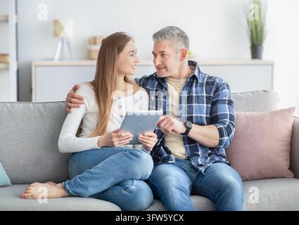 Happy mature couple with tablet pc sitting together on sofa at home, watching video, chatting with friend on webcam, checking email, shopping online, Stock Photo
