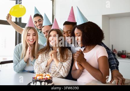 Young international friends congratulating Caucasian lady with her birthday, presenting festive cake, making wish before blowing candles, wearing part Stock Photo