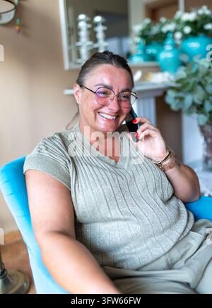 Cheerful woman smiling during a mobile phone conversation while sitting comfortably in her living room. Stock Photo