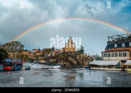 Tbilisi, Georgia - Apr 11, 2026: A full rainbow spans a stormy sky over Metekhi Church as a red tour bus and cars pass on a rain-soaked cobblestone st Stock Photo