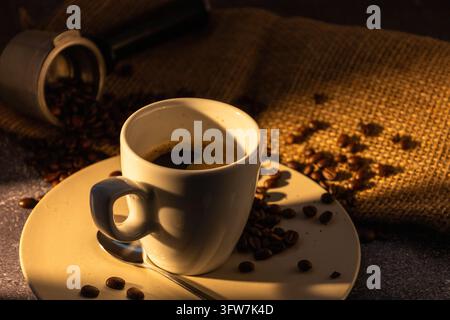 Steaming white cup of black coffee on a cream saucer with a spoon, surrounded by roasted coffee beans, a portafilter and burlap, in warm golden light Stock Photo