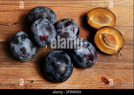 A top-down still life of ripe dark purple plums arranged on a rustic wooden surface, with two plums cut in half to reveal their golden-yellow flesh an Stock Photo