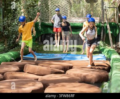 Group of friends have fun walking through an obstacle course next to a water in an amusement park, they are actively jumping on moving rocks Stock Photo