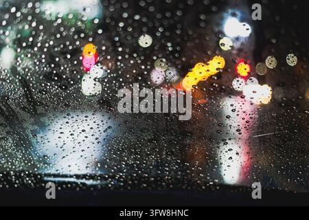 Raindrops on a car window with blurred colorful urban lights at night in Thailand, abstract rainy glass texture and bokeh background. Stock Photo