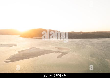 Aerial view of large sandbars in the shallow Sado estuary glowing in warm sunset light, Portugal. The sun sets behind the Arrabida hills on the left. Stock Photo