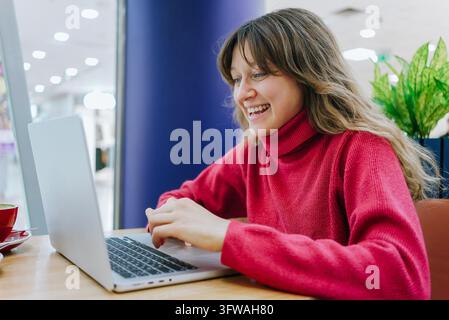 Happy female freelancer working on a laptop in a cozy cafe, savoring a cup of coffee while enjoying the relaxed atmosphere and natural light streaming through the window Stock Photo