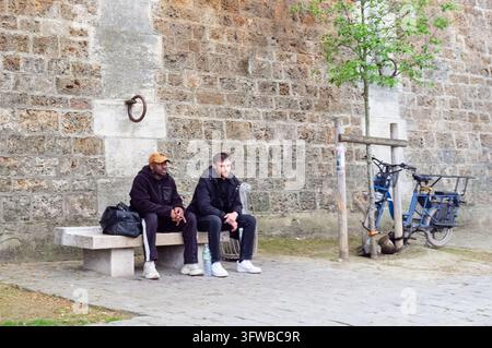 TWO MEN RELAXING ON A BENCH IN PARIS, SEINE LEFT BANK SUNDAY REPORTAGE, PARIS, FRANCE, 2026: Two young men, one wearing a baseball cap, sit together in thought on a concrete bench against an old stone wall, with a bicycle parked beside them. INFO: The Left Bank east of Notre-Dame blends academic heritage with riverside romance. Strolling past historic universities, tranquil botanical gardens, and the modern Arab World Institute reveals a vibrant, less-touristy pocket of Paris pulsing with intellectual soul. Stock Photo