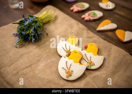 Handmade iced cookies shaped like chicks and eggs on rustic linen table with spring floral bouquet Stock Photo