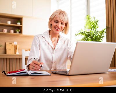 Positive remote professional smiling while working on a laptop beside her notebook and smartphone. Stock Photo