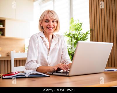 Smiling woman working on a laptop at her desk in a bright and comfortable home office. Stock Photo