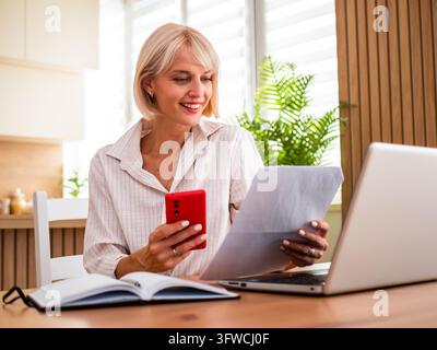 Smiling woman reviewing paperwork while using her smartphone and laptop at home. Stock Photo