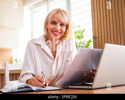 Happy businesswoman organizing paperwork and making notes during a productive workday at home. Stock Photo