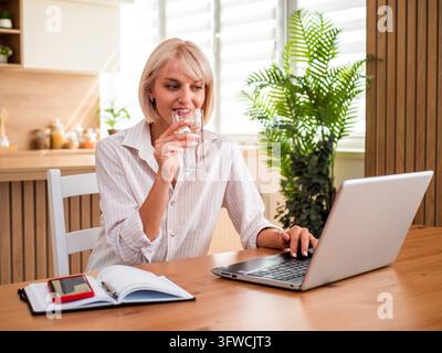 Blond woman working from home, typing on laptop and drinking water. Stock Photo