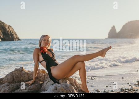 Blonde adult female wearing black swimsuit posing on coastal rock formation near ocean waves during golden hour Stock Photo