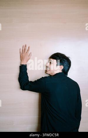A young man wearing a black shirt touches a light brown wooden wall with his hand raised, posing indoors with soft lighting. Stock Photo