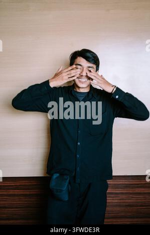 A young adult man wearing a black shirt stands indoors against a wooden wall, smiling with his eyes covered by his hands. Stock Photo