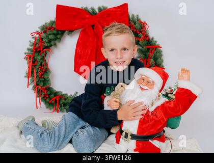 Cute boy hugging plush Santa toy sitting on floor during Christmas photoshoot in white studio. Big decorated spruce wreath, festive sweater, children Stock Photo