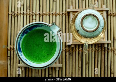 A serene overhead composition of freshly whisked matcha green tea in a ceramic bowl alongside a glass teapot arranged on a rustic bamboo surface Stock Photo
