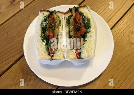 A sandwich cut in half, filled with meat, greens, and sauce, served on a white plate on a wooden table. Stock Photo