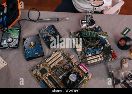 Top view of technician workspace with various computer components like motherboards, hard drives RAM sticks. Soldering iron and other tools suggest Stock Photo