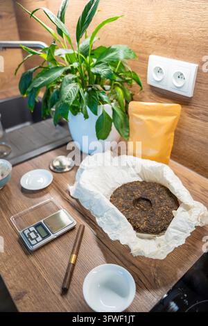 A compressed puer tea cake lies on paper beside a digital scale and tea pick. Measuring loose portions prepares Chinese tea for home brewing. Stock Photo