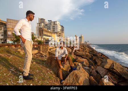 Two young men chat on the rocks by the sea near city buildings, Western Province, Colombo, Sri Lanka Stock Photo