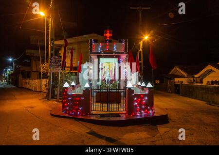 Illuminated Christian roadside shrine with red flags at night on an empty street, Western Province, Negombo, Sri Lanka Stock Photo