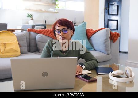 Woman typing on silver laptop at glass coffee table with purple smartphone, wearing green top Stock Photo