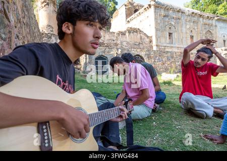 Young man playing an acoustic guitar with friends sitting on grass near old ruins, Jammu and Kashmir, Srinagar, India Stock Photo