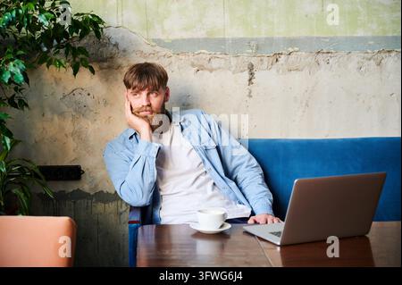 Thoughtful bearded man sitting with laptop and coffee in cafe interior, resting face on hand while looking away in quiet remote work moment Stock Photo
