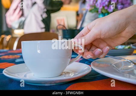 White ceramic coffee cup on a saucer with spoon, held by a manicured hand at a colorful cafe table beside flowers and a plat Stock Photo