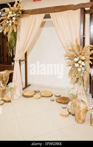 Rustic wedding ceremony arch decorated with dried pampas grass bouquets and beige curtains, wooden stumps on tiled floor Stock Photo
